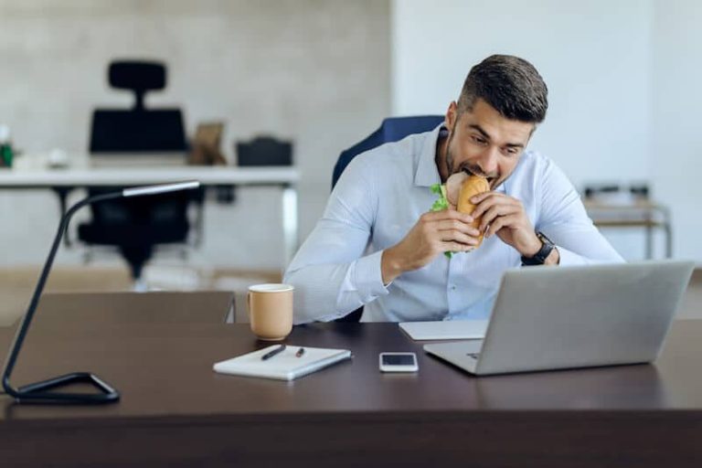Un individu déguste un sandwich tout en travaillant devant son ordinateur au bureau.