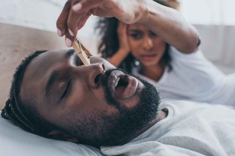 Homme qui ronfle bruyamment et sa femme lui place une pince sur le nez pour stopper le bruit.
