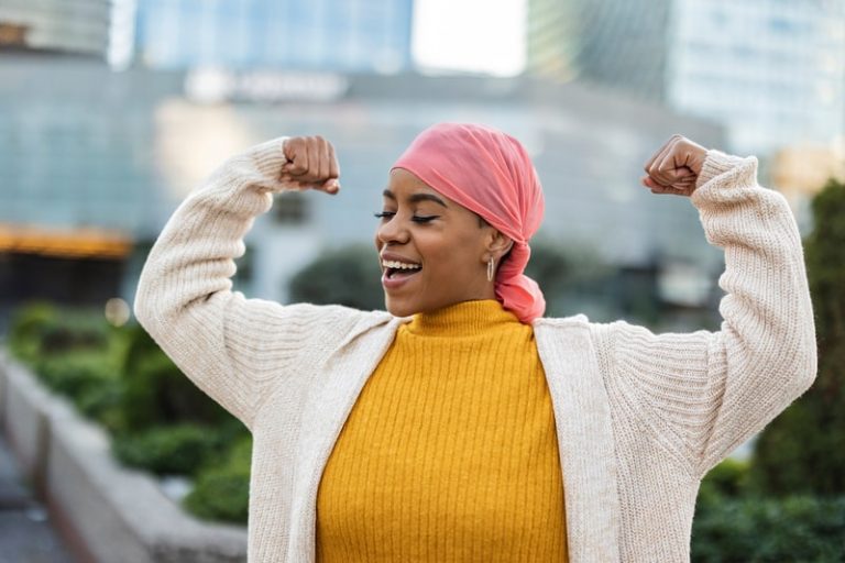 Une femme qui lutte contre le cancer du sein, porte un foulard rose et lève les bras comme une combattante