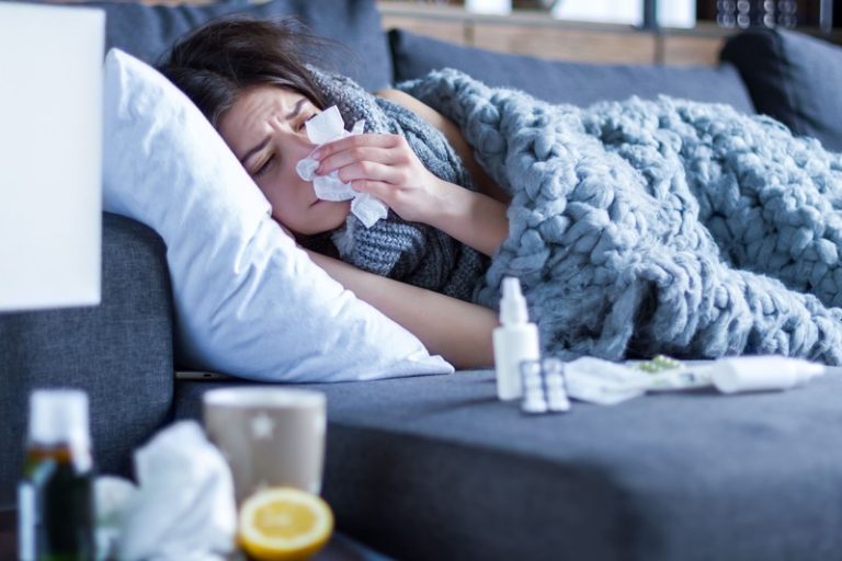Une femme malade à cause d'un virus hivernal, allongée sur son canapé avec médicaments et mouchoirs.