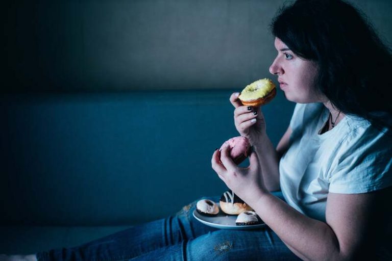 Une femme sur son canapé, dans le noir, en dépression, en train de consommer des aliments remplis de sucre.