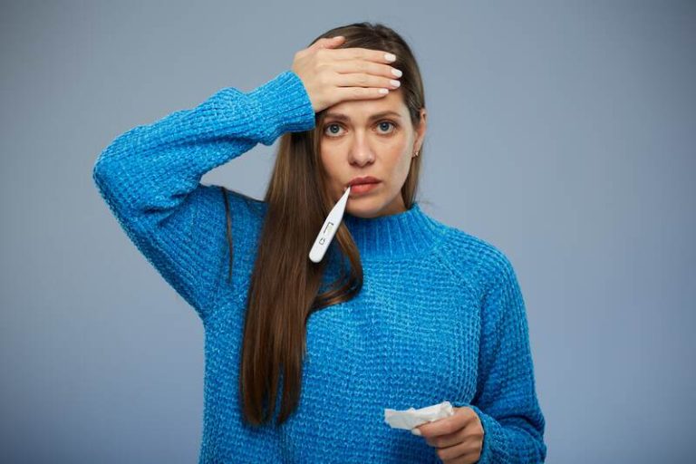 Une femme souffrant des symptômes du covid long avec un thermomètre dans la bouche et la main sur le front.