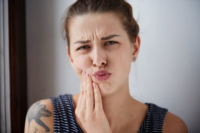 Une femme se tord de douleur en se tenant la joue, gênée par un aphte douloureux dans la bouche.