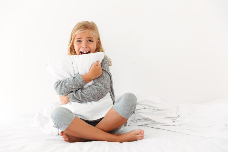 Jeune fille assise sur le lit, serrant un oreiller contre elle avec une expression inquiète.