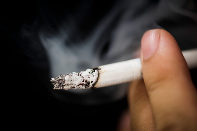 Une personne tient une cigarette allumée, dont la fumée se diffuse dans l’air et expose les autres au tabagisme passif.