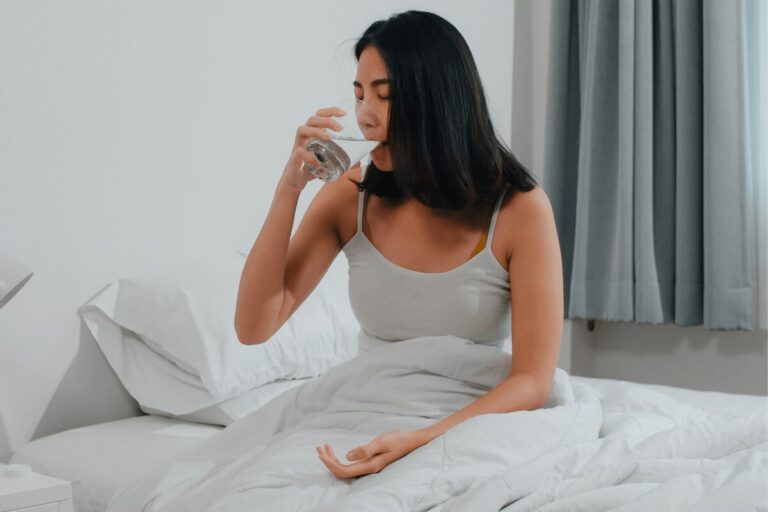 Femme buvant un verre d’eau dans son lit au réveil, suspectant une xérostomie en raison d’une sensation de bouche sèche.