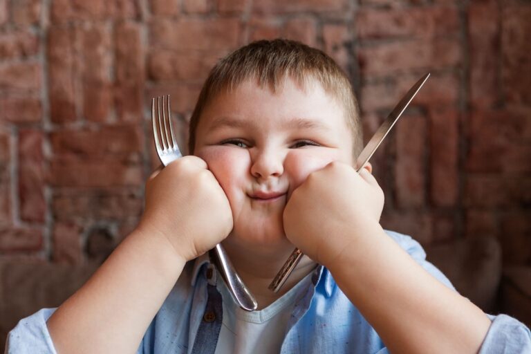 Un enfant présentant un début d’obésité attend son repas, les couverts à la main.