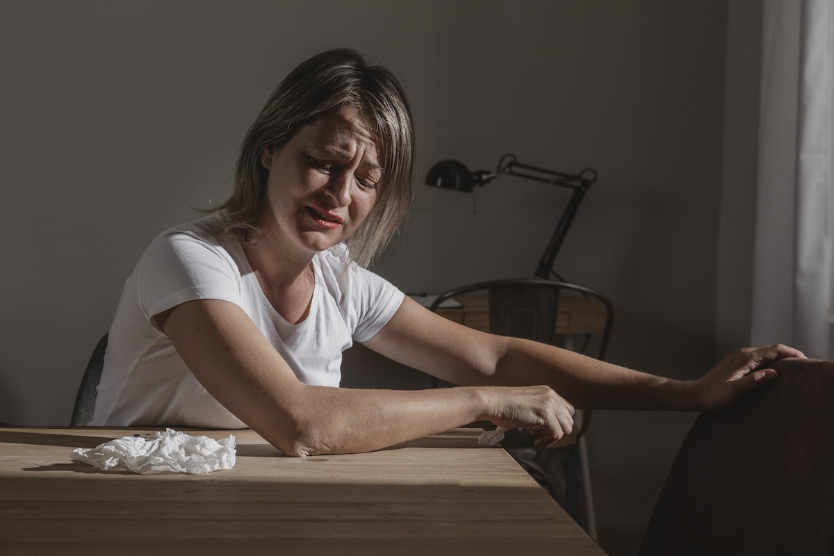 Une femme assise à une table, le visage crispé et les yeux fermés, semble souffrir de douleurs chroniques qui l'épuisent.