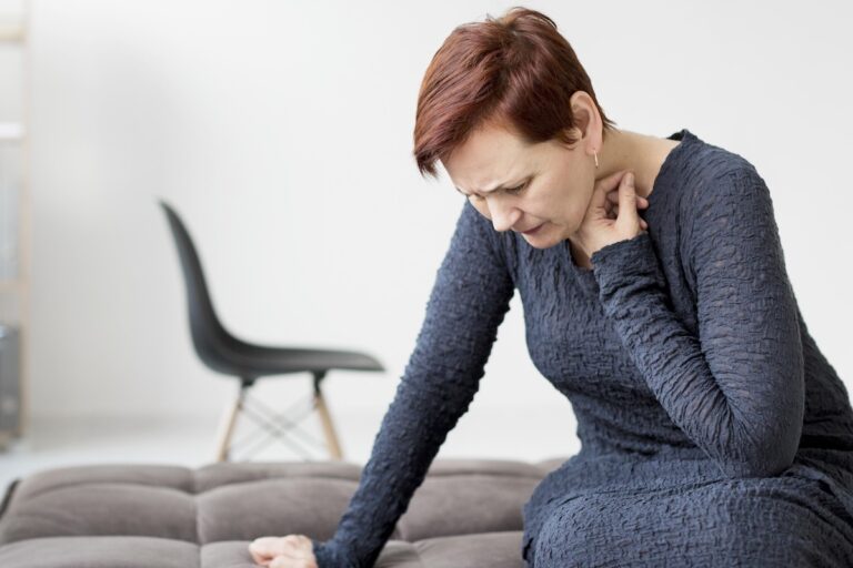 Une femme assise sur un canapé se tient le cou, le visage crispé, semblant souffrir d’une douleur cervicale.