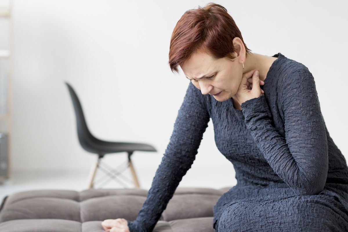 Une femme assise sur un canapé se tient le cou, le visage crispé, semblant souffrir d’une douleur cervicale.