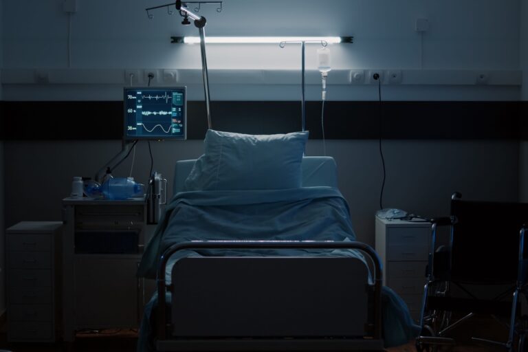 Un lit d'hôpital vide, entouré d'équipements médicaux, dans une chambre plongée dans la pénombre.