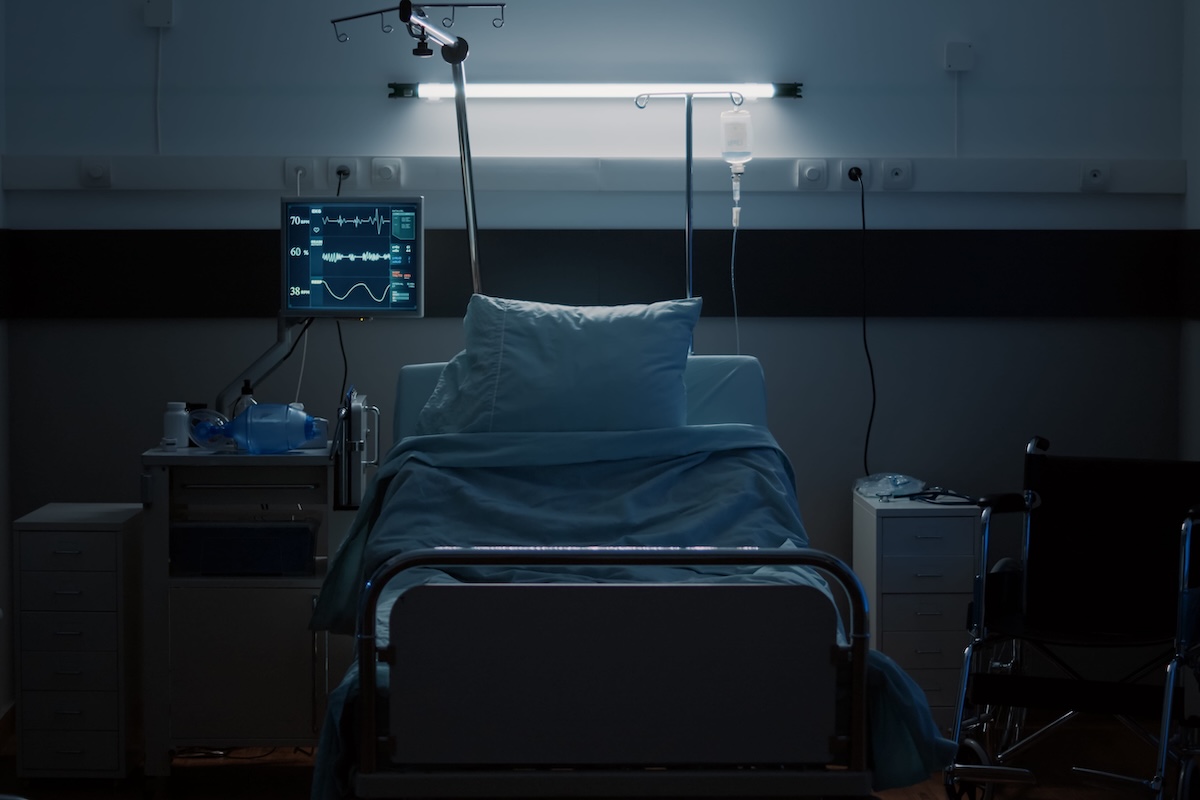 Un lit d'hôpital vide, entouré d'équipements médicaux, dans une chambre plongée dans la pénombre.