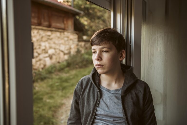 Un adolescent regarde par la fenêtre, l’air triste et pensif, assis seul dans une pièce.