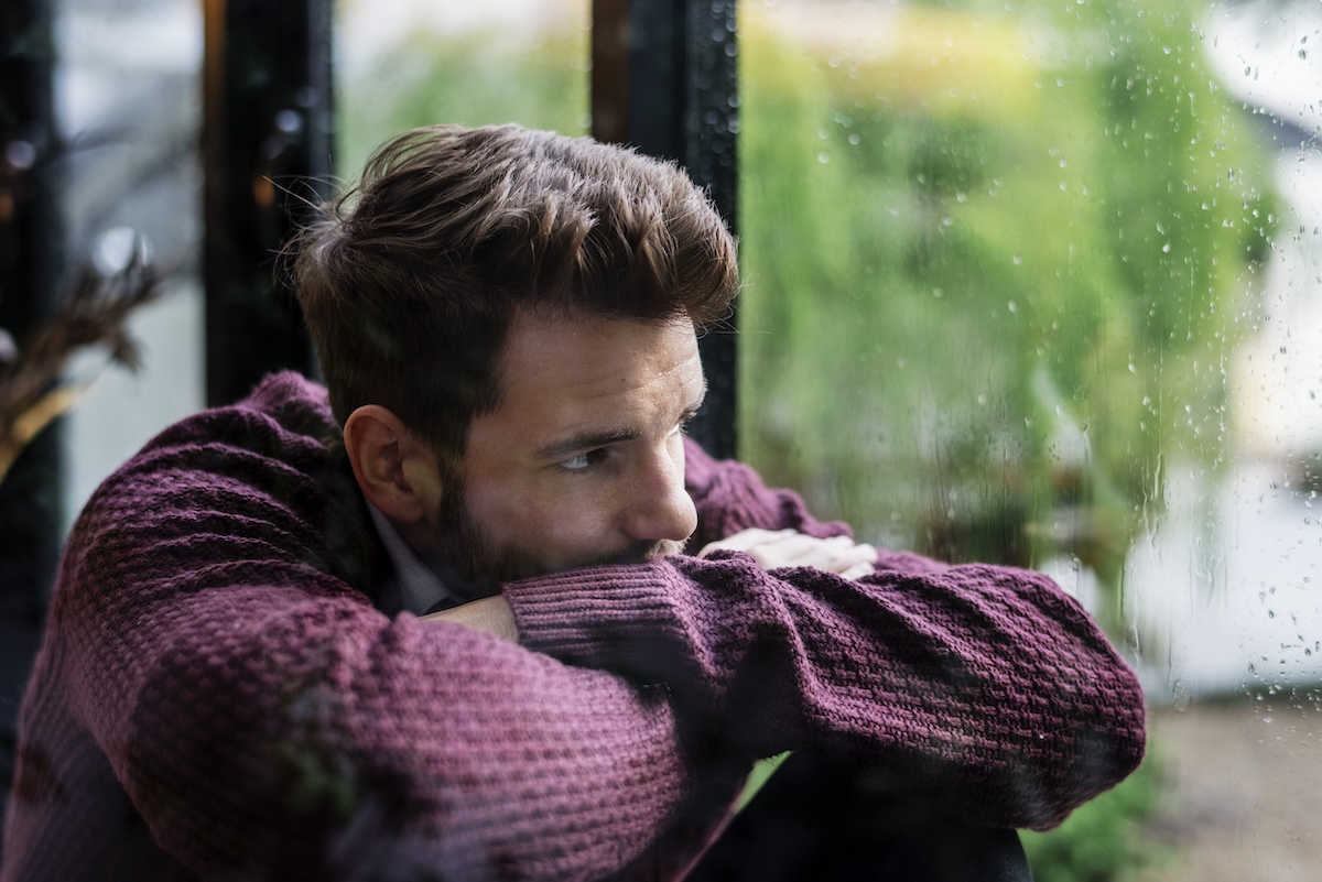 Un jeune homme, l’air triste et pensif, est assis les bras croisés, le regard perdu à travers une fenêtre.