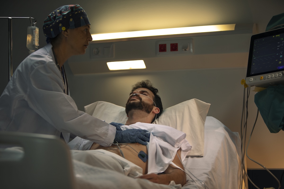Un médecin examine un patient contaminé par le virus West Nile, allongé sur un lit d’hôpital, sous surveillance cardiaque et assistance en oxygène, dans une unité de soins intensifs.