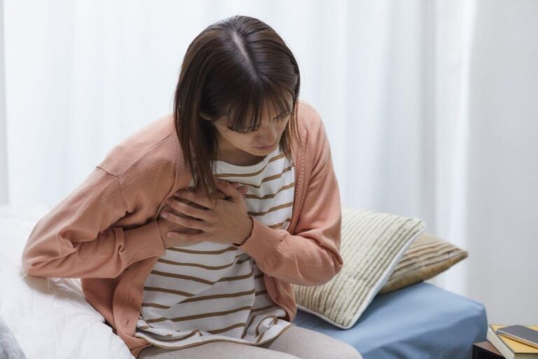 Une femme assise sur un lit se penche vers l’avant en se tenant la poitrine des deux mains, suspectant une douleur thoracique liée à une péricardite.